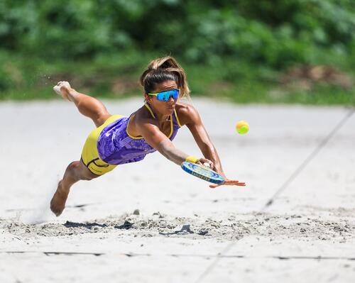 Eine Frau springt mit einem Beachtennisschläger in der Hand nach vorne in den Sand.