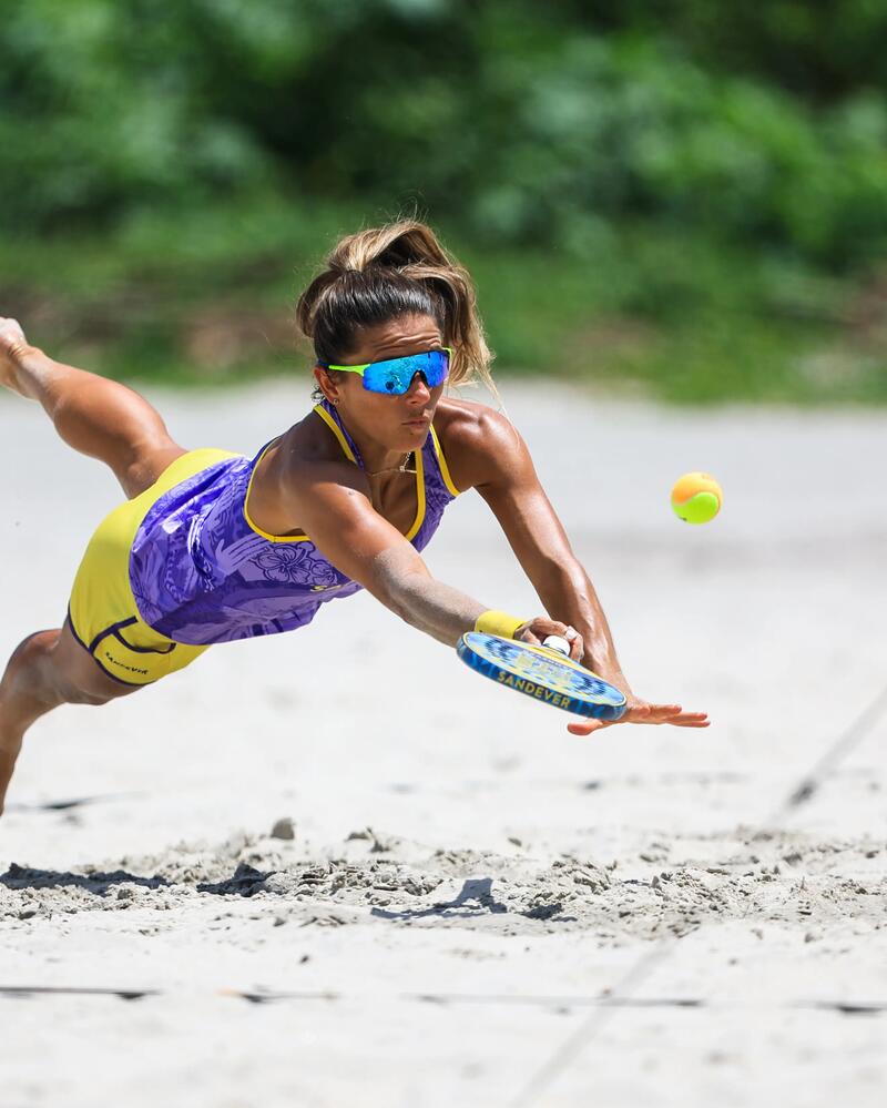Ein Vater hilft seiner Tochter am sonnigen Strand dabei, den Beach-Tennis-Schläger richtig zu halten, während ihnen eine weitere Person einen Ball zuspielt.
