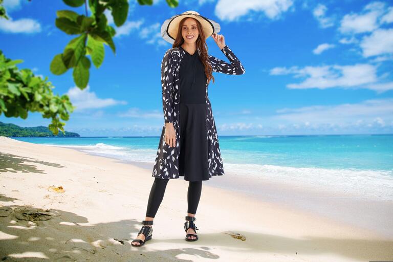 Woman wearing a black floral swim dress with leggings and a sun hat on the beach.