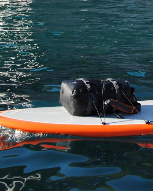 Orange-weißes Surfbrett schwimmt auf blauem Wasser, schwarze Tasche darauf befestigt, glänzende Wellenoberfläche.