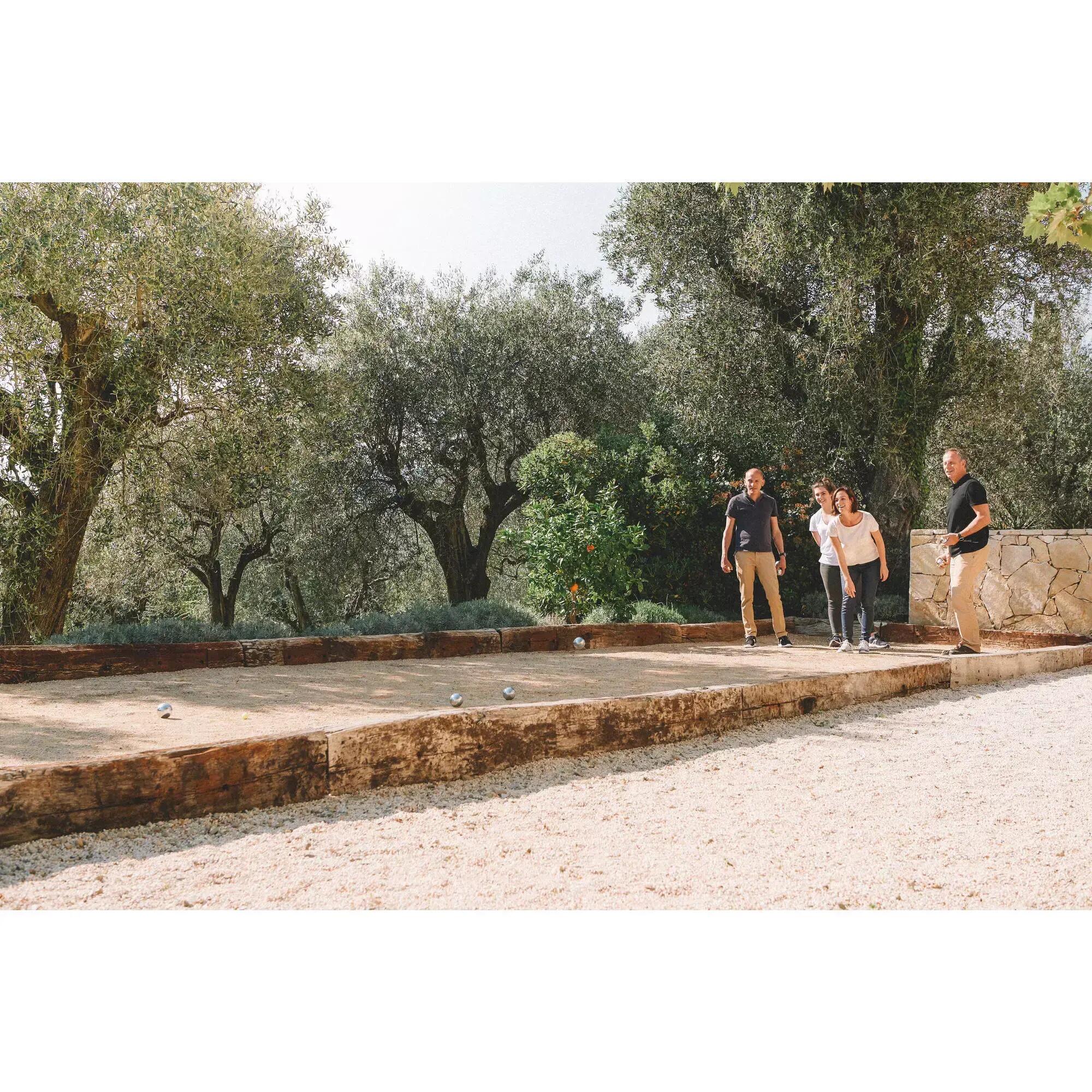 Eine Gruppe von Erwachsenen spielt Pétanque auf einer Sandbahn im Freien umgeben von Bäumen.
