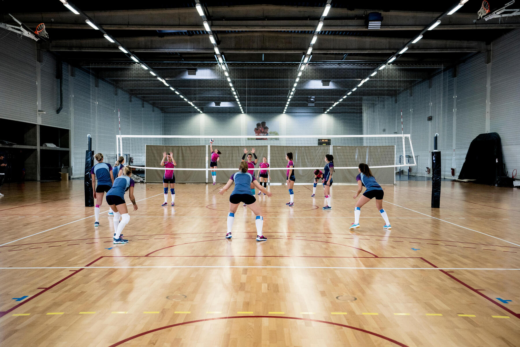 Zawodniczki w strojach siatkarskich ustawione na boisku przygotowane do odbioru piłki od siatkarki wykonującej serwis podczas meczu siatkówki żeńskiej na hali sportowej