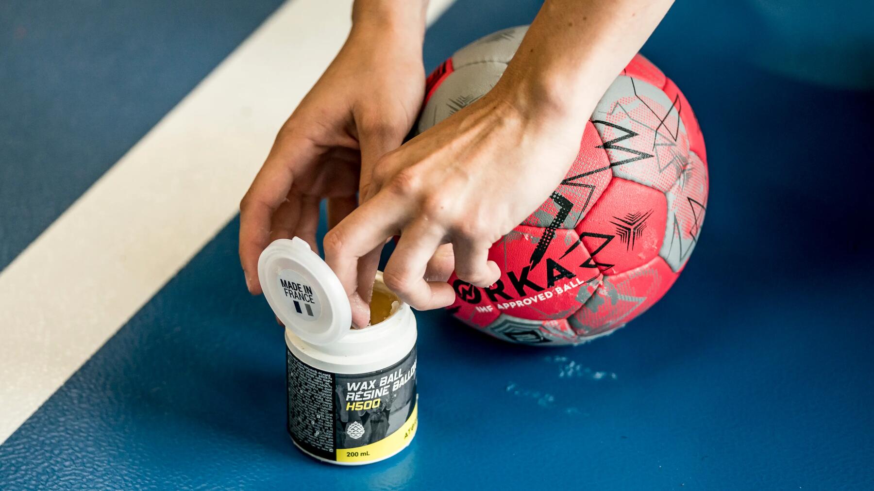 Hände öffnen Handballwachs neben rotem Handball, blaue Spielfläche, sportliche Vorbereitung.