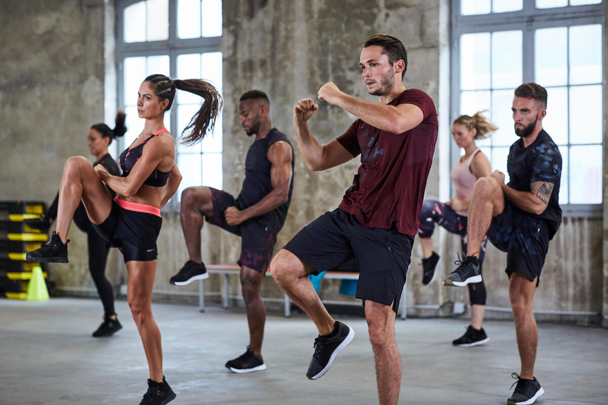 Skupina moških in žensk v športnih oblačilih izvaja intenziven aerobni trening v prostorni dvorani industrijskega stila.