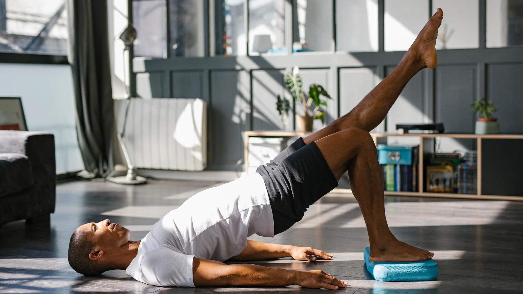 Mann in weißem Sportshirt und grauer kurzer Sporthose auf dem Boden mit blauem Yogablock beim Pilates