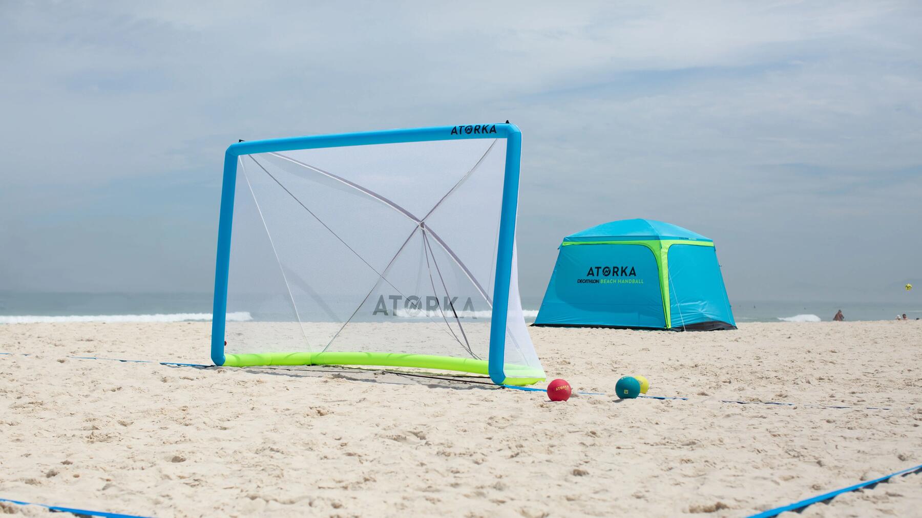 Frau in blauer Sportkleidung wirft Ball am Strand, Mann in schwarzem Top verteidigt, Beachhandballspiel.