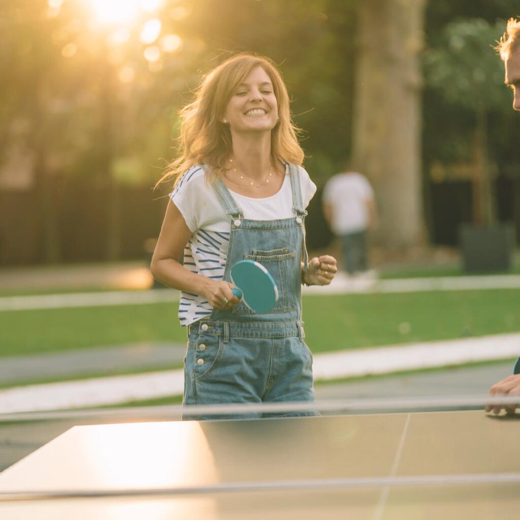 Eine lachende Frau in Latzhose und ein Mann spielen im Gegenlicht der Abendsonne Tischtennis im Park.