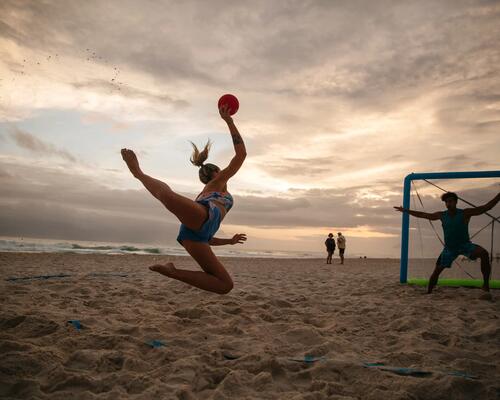 Person springt mit Ball am Strand, Sonnenuntergang im Hintergrund, Beach-Handball-Szene, wolkiger Himmel, sportliche Dynamik.