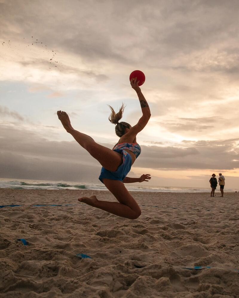 Person springt mit Ball am Strand, Sonnenuntergang im Hintergrund, Beach-Handball-Szene, wolkiger Himmel, sportliche Dynamik.