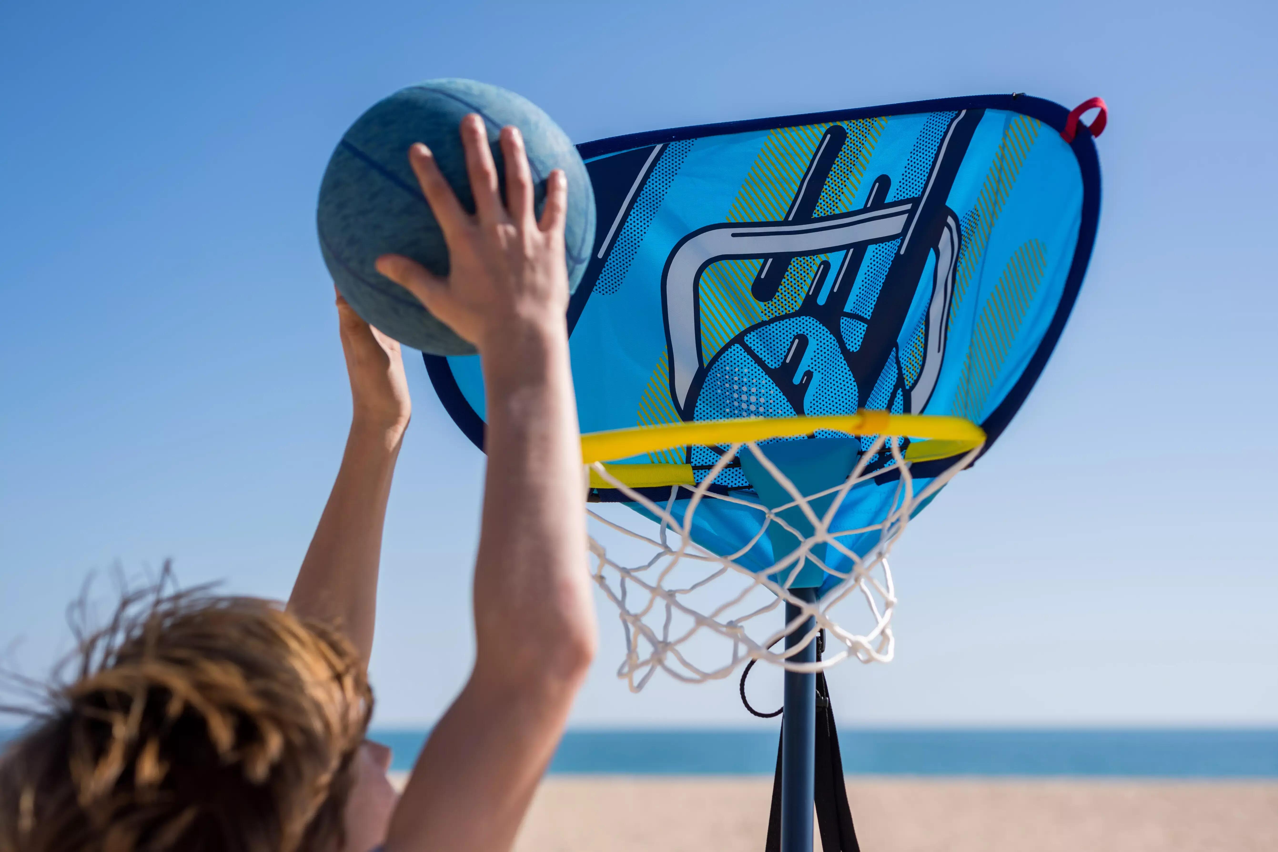 Child shooting basketball into blue hoop, close-up, ocean in background