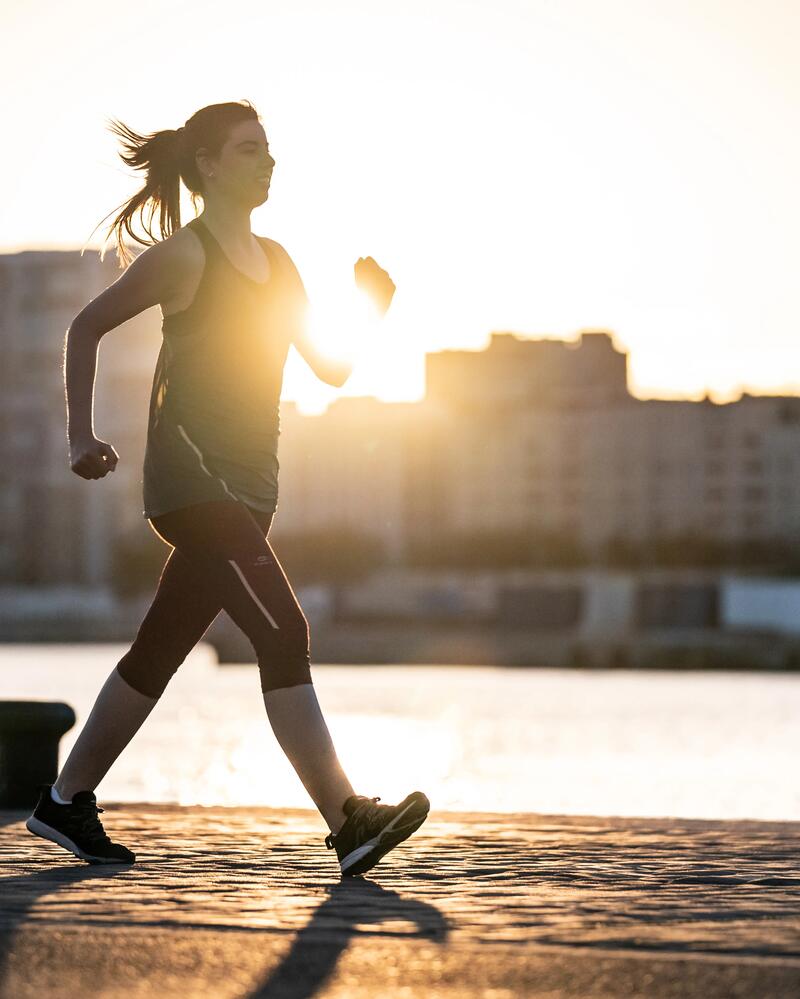 Frau geht zügig auf einer Uferpromenade vor einer städtischen Silhouette im Gegenlicht der Abendsonne.