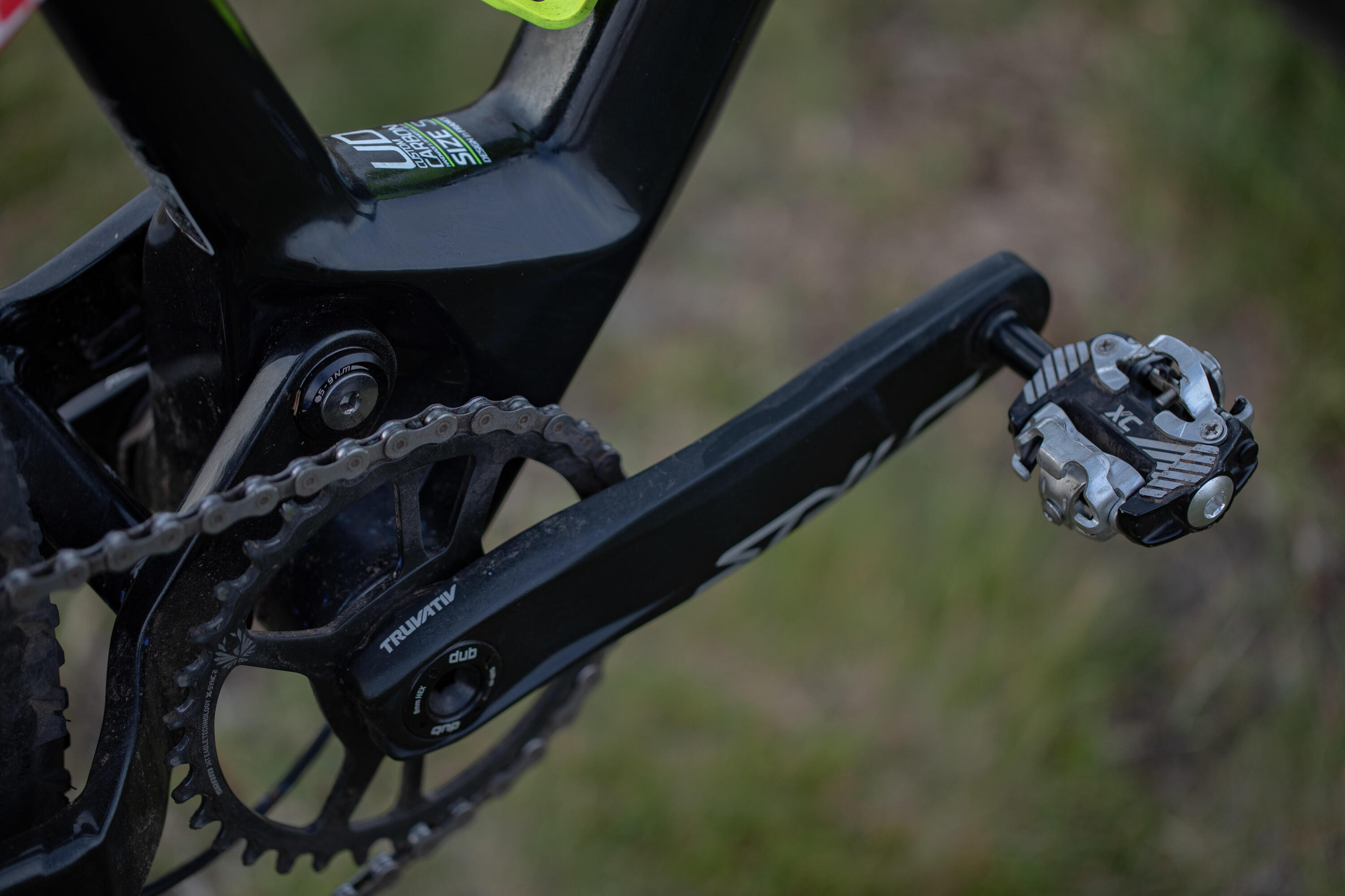 Eine Nahaufnahme des schwarzen Kurbelarms eines Fahrrads mit einem montierten Klickpedal sowie Kette und Kettenblatt. Im Hintergrund ist eine unscharfe Grasfläche zu sehen.