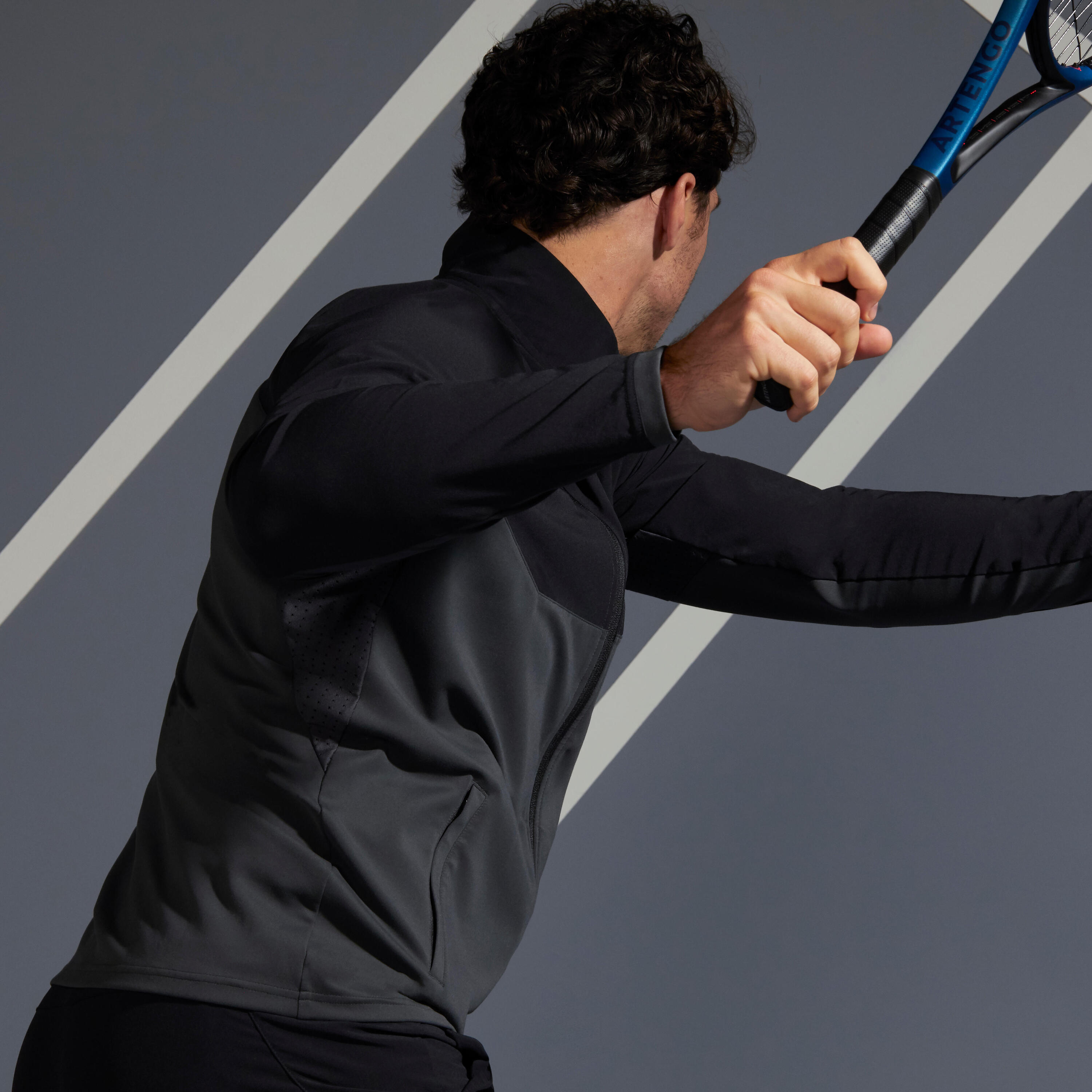 Rückansicht eines Mannes in dunkler Sportjacke, der dynamisch mit einem Artengo-Tennisschläger ausholt.