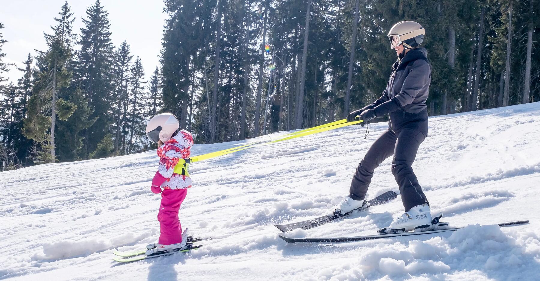 A girl and her daughter going down the slope on the skis and the using a harness help the girl ski