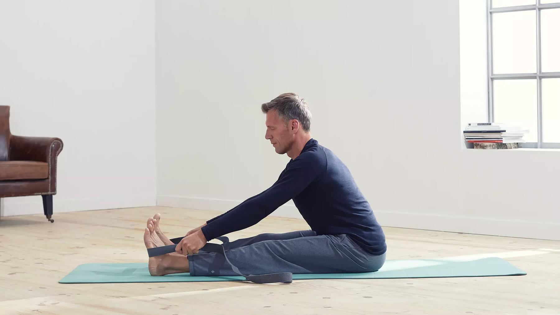 Mann in blauer Yoga Bekleidung auf türkiser Yogamatte mit Yogagurt in den Händen bei Übung.