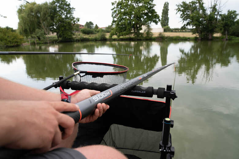 Ribič drži dolgo 9-metrsko ribiško palico Caperlan Carpower nad vodo, v ozadju je viden podmetač za ribe.