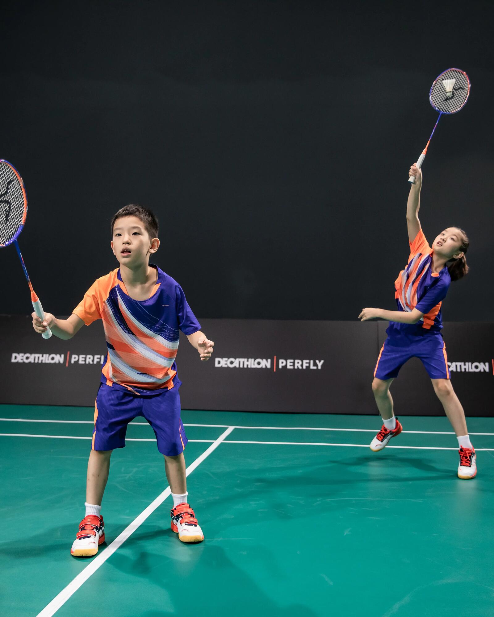 Zwei Kinder spielen Badminton in lila-orangefarbener Sportkleidung auf einem grünen Spielfeld.