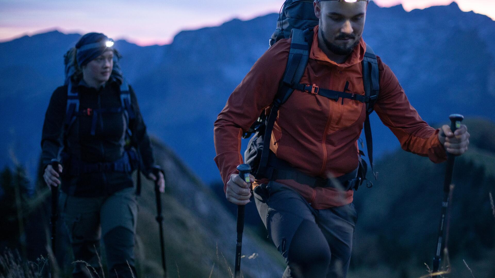 mężczyzna i kobieta idący po górach z kijami trekkingowymi w rękach i latarkami czołowymi na głowach