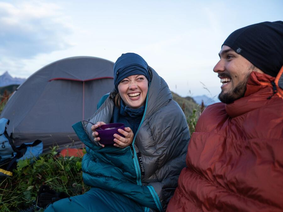 man and woman sat eating by their campsite the woman wears a sleeping bag for warmth