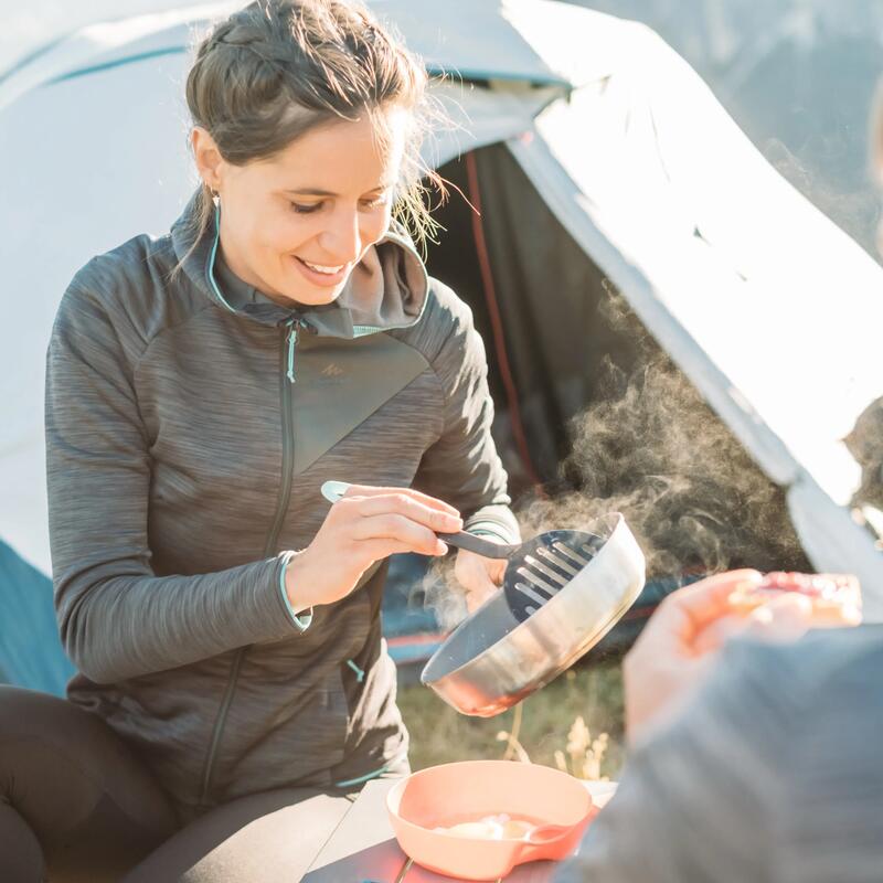 Frau sitzt vor Zelt und ist mit Camping- Kochequipment am kochen.