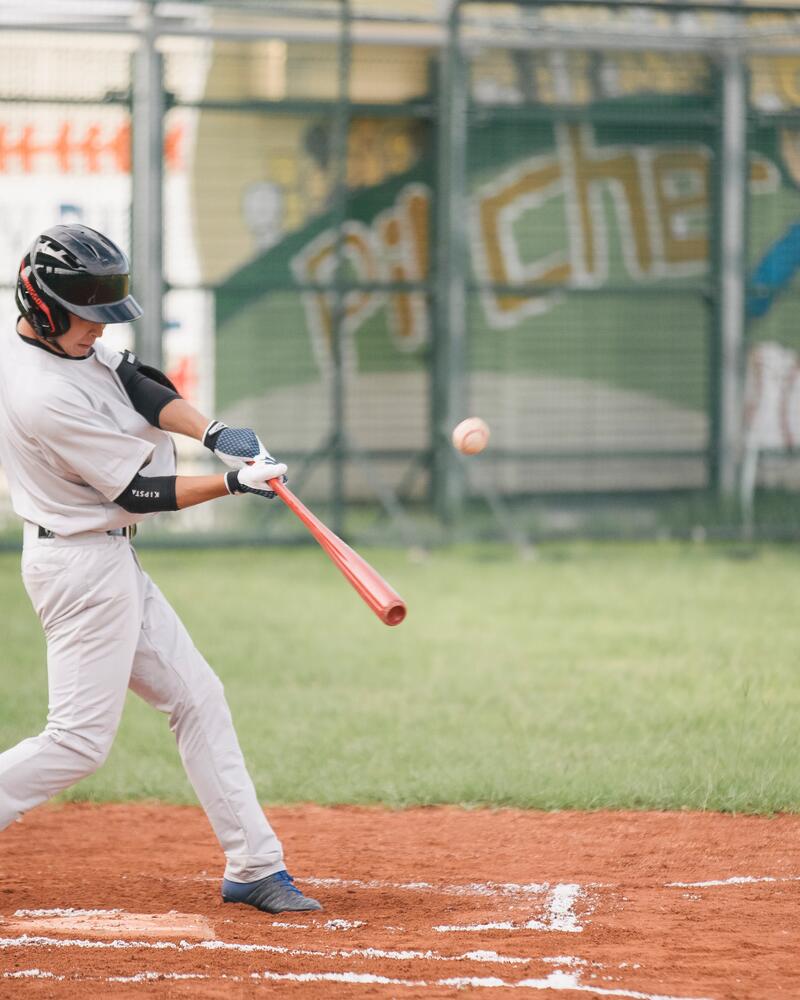 Ein Baseballspieler in hellgrauer Uniform und schwarzem Helm schwingt auf einem rötlichen Spielfeld kraftvoll seinen roten Schläger, um den herannahenden Ball zu treffen.