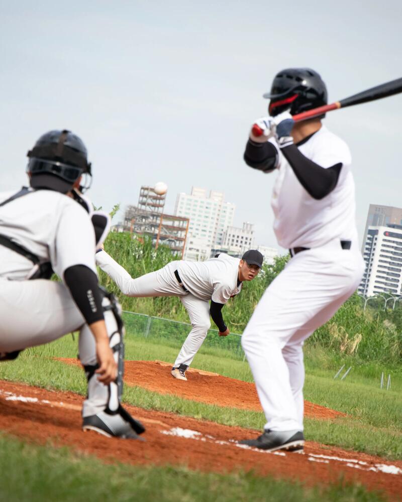 Ein Baseballspieler schlägt an der Home Plate einen Ball, der gerade vom Pitcher auf dem Wurfhügel geworfen wurde, während der Catcher im Vordergrund in der Hocke wartet.