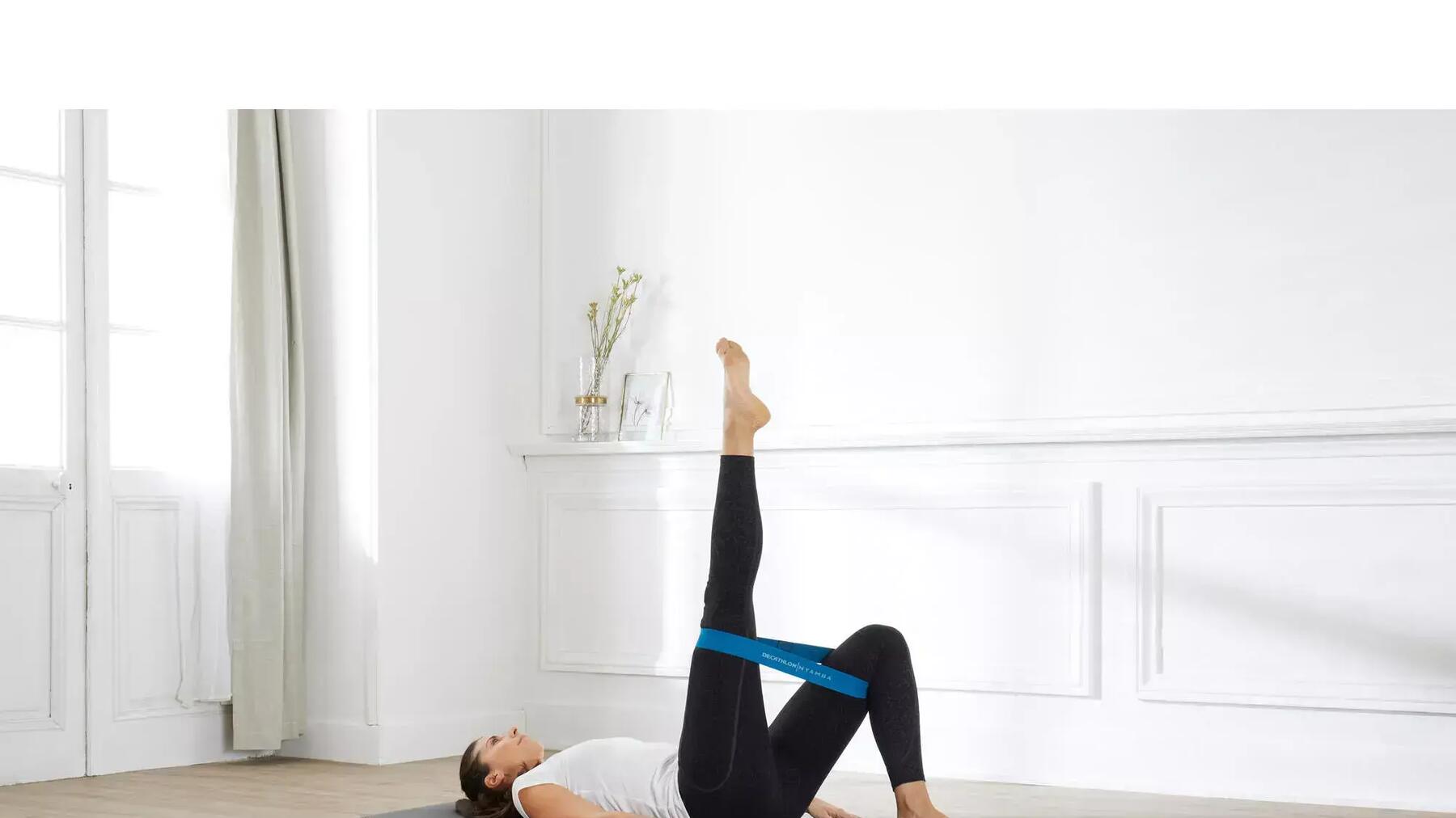 Frau auf grauer Yogamatte in schwarzer Leggings und weißem Sportshirt mit blauem Band um die Oberschenkel beim Pilates