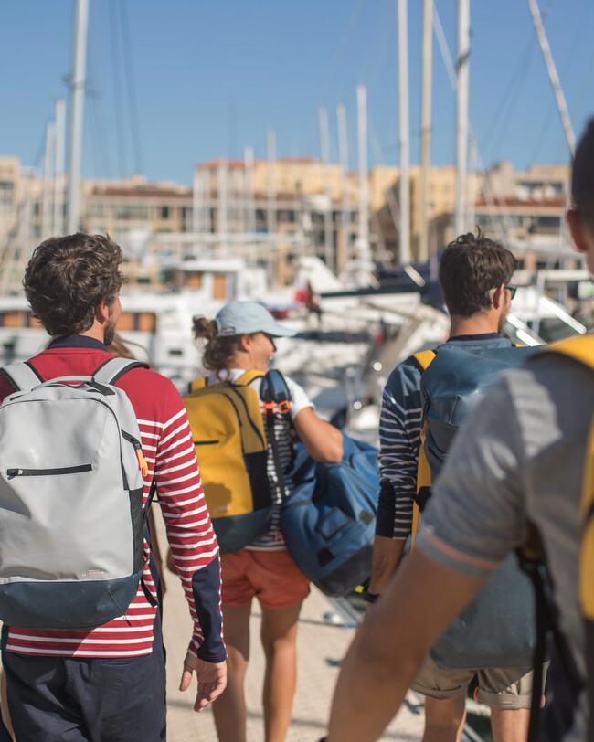 Personen mit bunten Rucksäcken spazieren an einem sonnigen Hafen entlang, umgeben von Booten und Masten.
