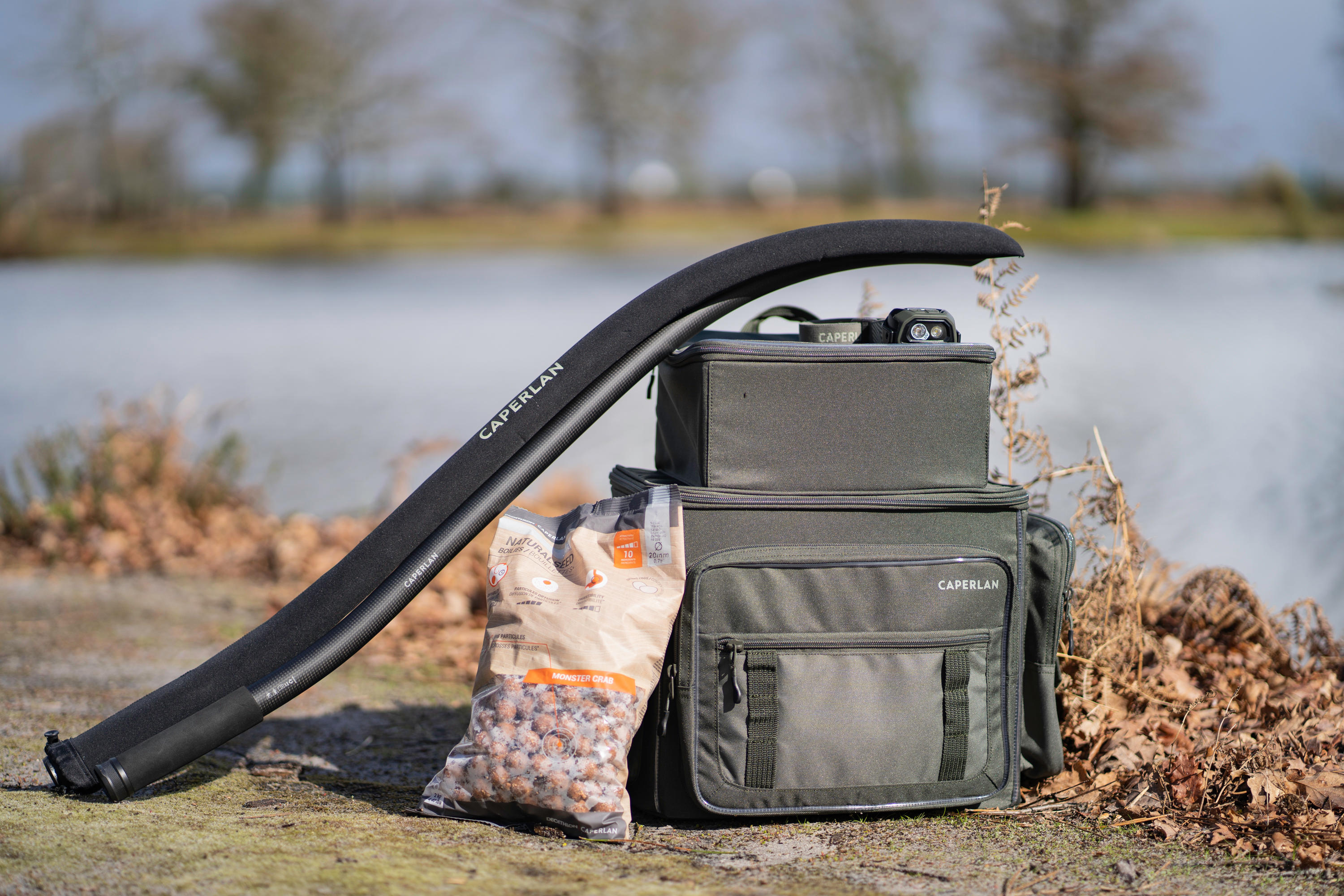 Eine Angeltasche mit einer Packung Boilies und einem Futterrohr stehen auf dem Boden am Wasser