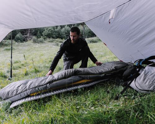 mężczyzna rozkładający śpiwór turystyczny na karimacie trekkingowej pod rozłożonym namiotem