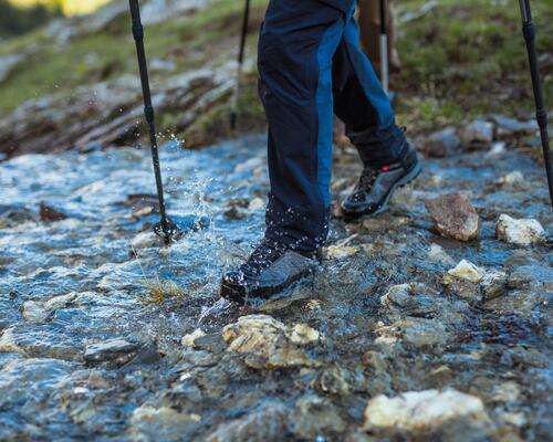 mężczyzna idący w butach trekkingowych z kijkami przez strumyk 