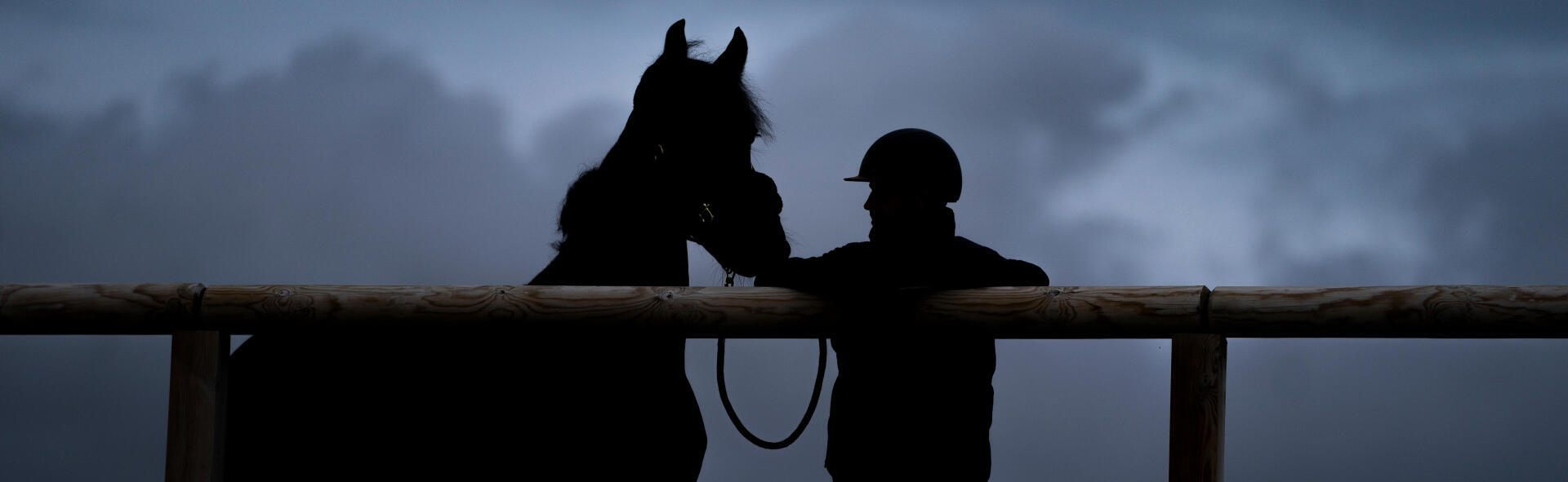 Un cavalier s'appuie sur la barrière en bois de sa carrière gelée, son cheval en longe à côté de lui