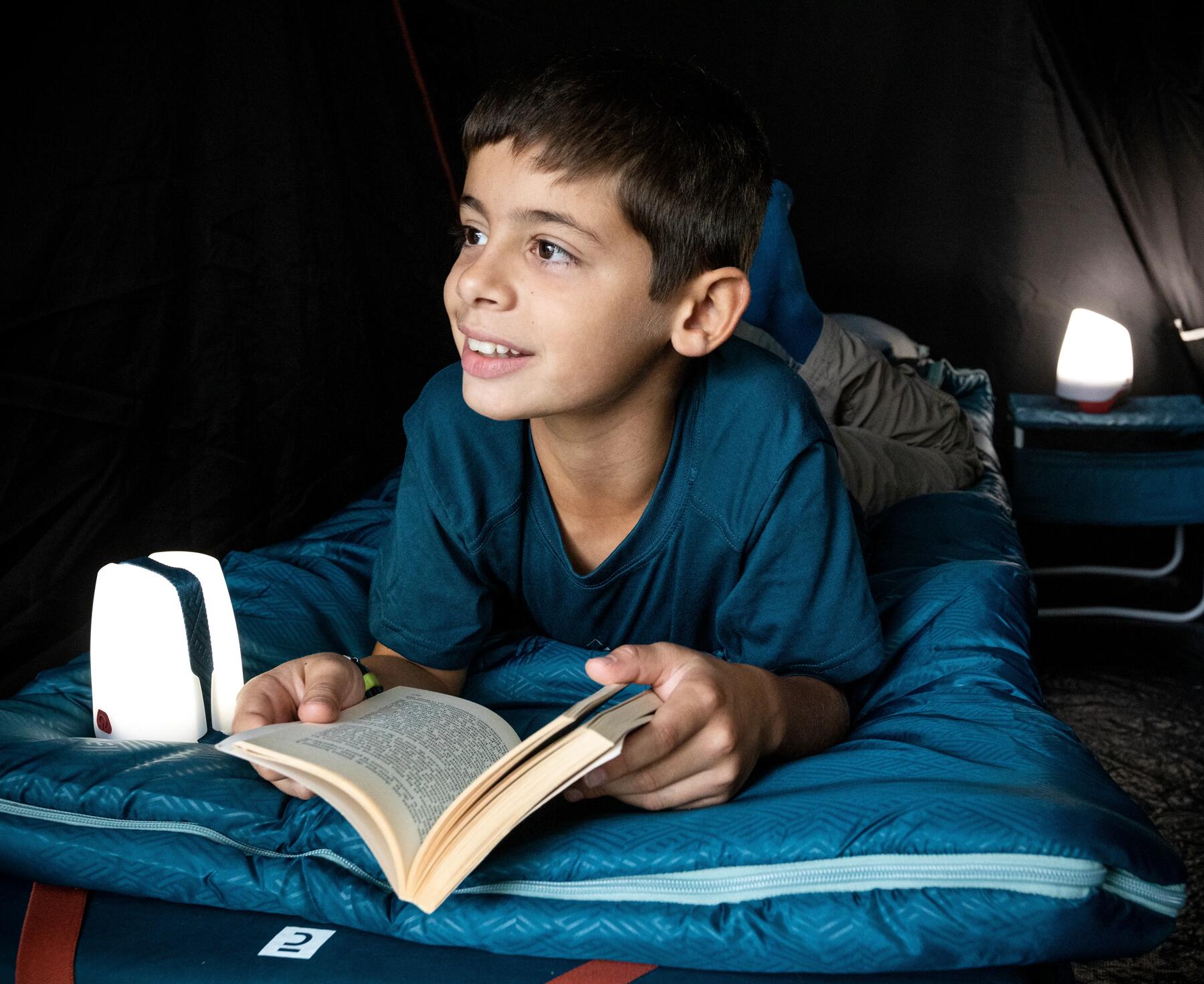 Boy lays up on a sleeping bag reading a book with a camp light next to him