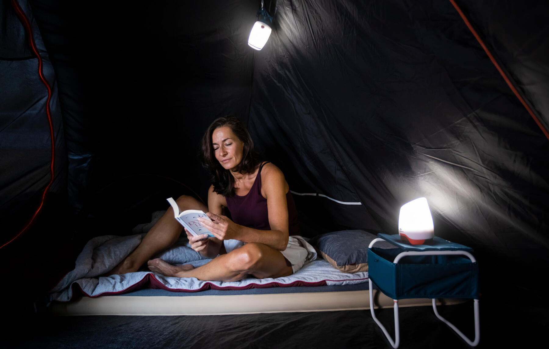 A woman reads comfortably in a tent with a mattress, sleeping bag, and light