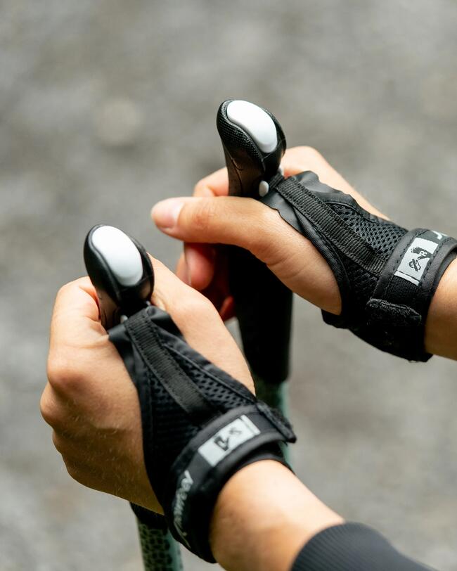Hände halten schwarze Nordic-Walking-Stöcke, Erdton-Hintergrund, leuchtendes Armband, Fokus auf Griffe und Textur der Handschuhe.
