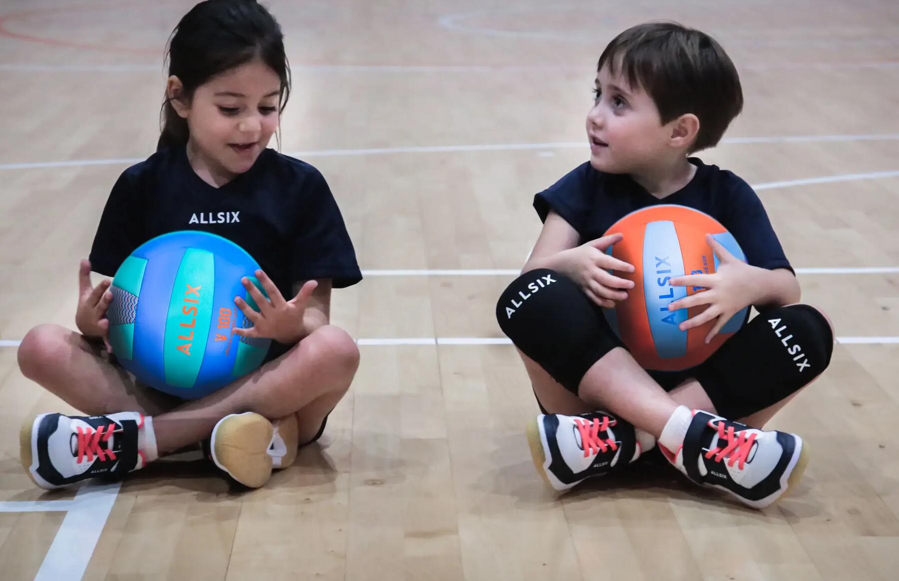 Zwei Kinder tragen Volleyballschuhe und sitzen in der Sporthalle auf dem Boden