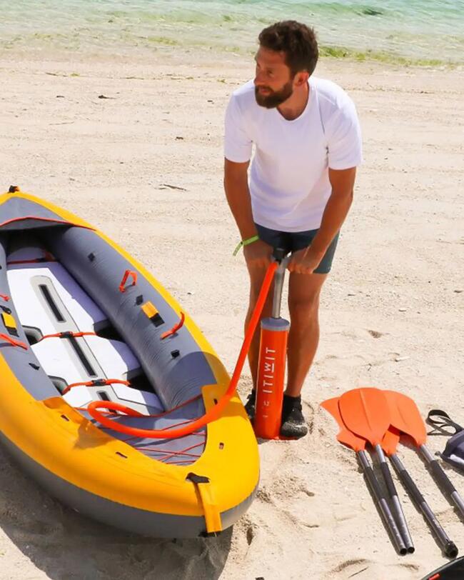 Mann pumpt gelbes Schlauchboot auf, orange Paddel im Sand, blauer Himmel, sonniger Strand, klare Formen und farbenfrohe Szenerie.
