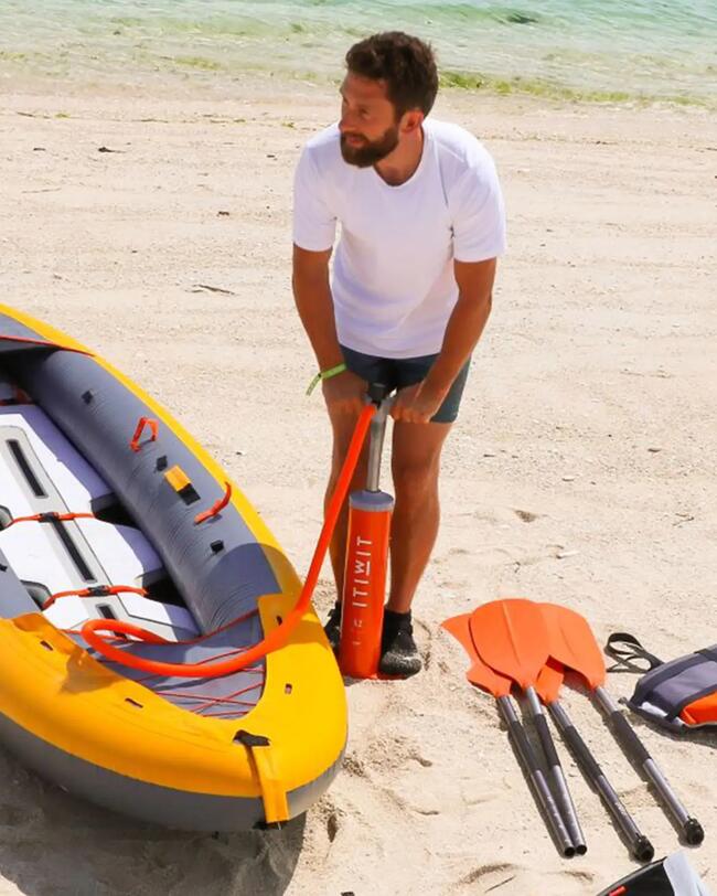 Mann pumpt gelbes Schlauchboot am Strand auf, orangefarbene Paddel, weißer Sand und blaues Wasser im Hintergrund.