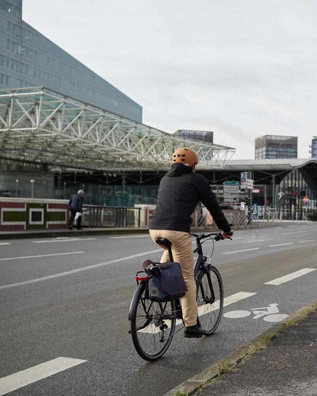 Mann mit orangem Helm fährt auf grauem Fahrrad mit Taschen eine Straße entlang. Im Hintergrund Gebäude