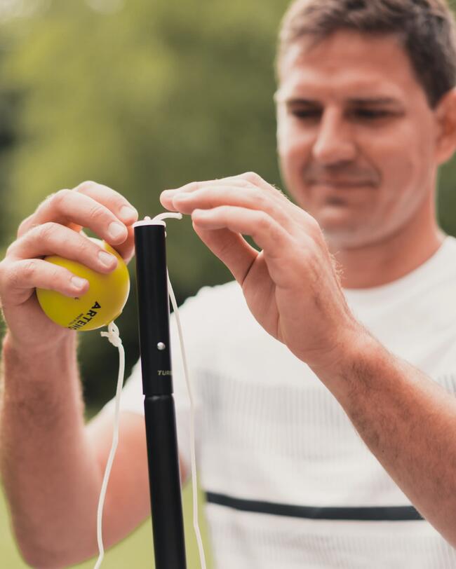Ein Mann in einem weißen T-Shirt befestigt mit seinen Händen einen leuchtend gelben Ball an dem schwarzen Masten des Speedball Sets.