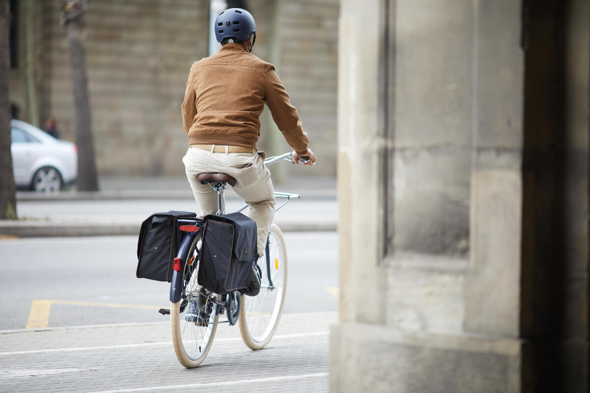 Biciklista sa braon jaknom i plavom kacigom vozi gradski bicikl sa crnim torbama.