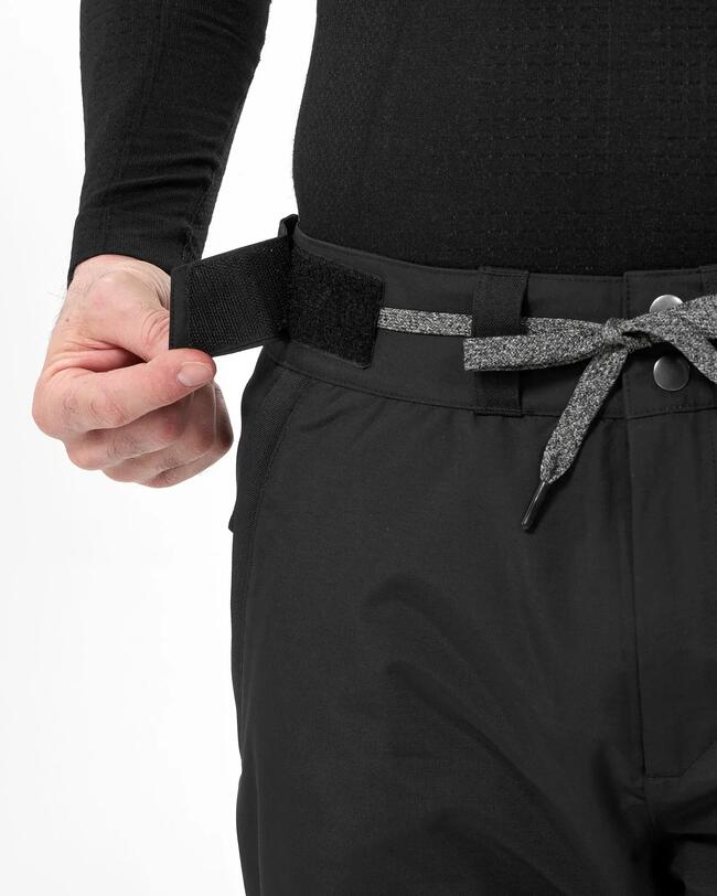 Ein Mann demonstriert mit seiner Hand den mit Klettverschluss verstellbaren Hosenbund der schwarzen Snowboardhose.