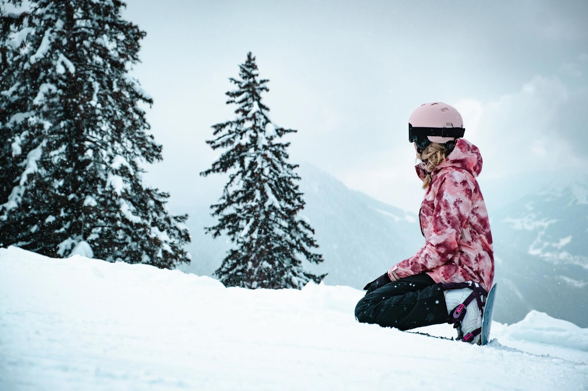 Kobieta w odzieży i kasku snowboardowym odpoczywająca na stoku z przypiętą deską snowboardową