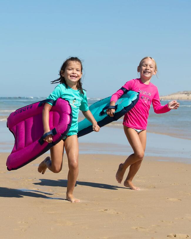 Zwei lachende Kinder in bunten Badeanzügen rennen am Strand mit pinken und blauen Surfbrettern.