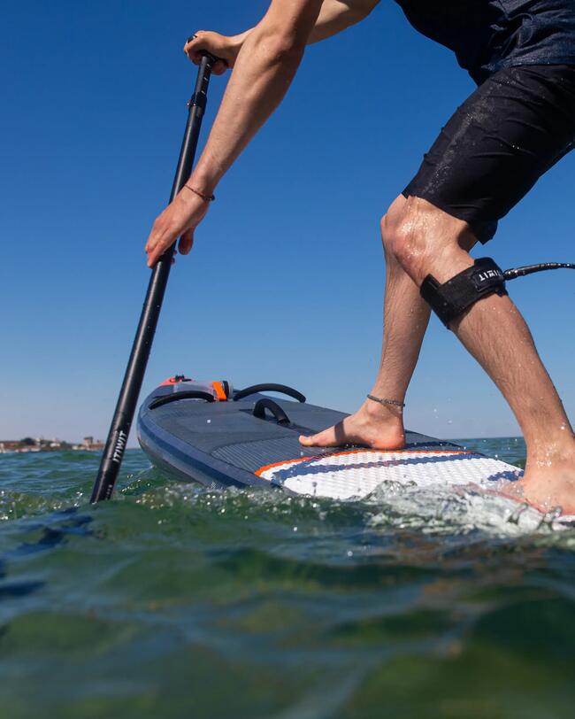 Person paddelt auf einem blauen SUP-Board, klarer Himmel, grünes Wasser, Küste im Hintergrund.