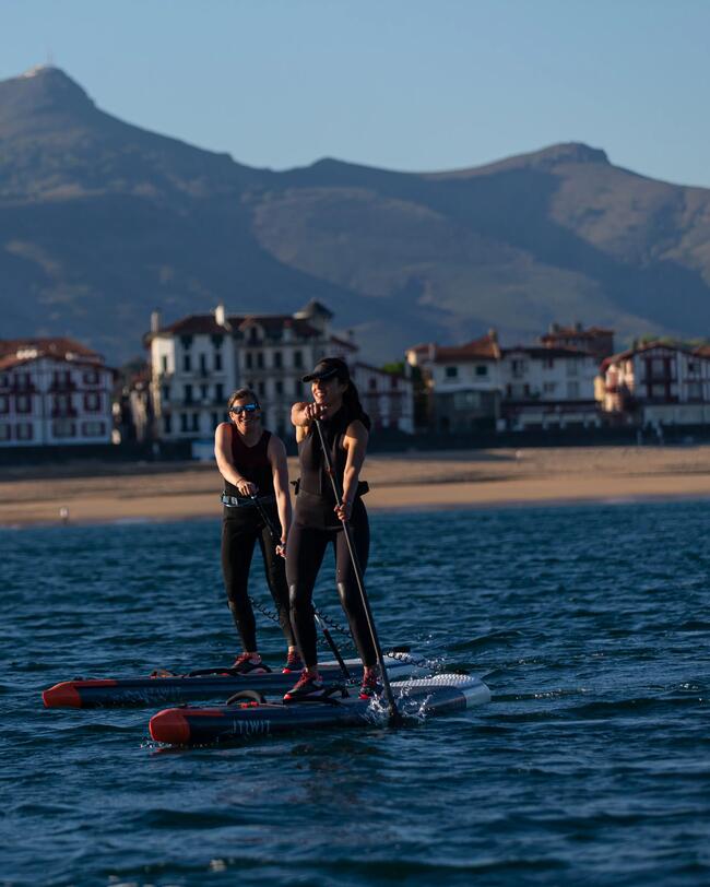 Menschen auf Paddleboards im blauen Wasser, im Hintergrund Häuser, Sandstrand und Berge unter klarem Himmel.