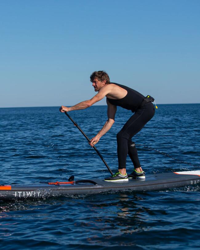 Mann auf Paddleboard, dunkelblauer Neoprenanzug, sonniger Himmel, ruhiges blaues Wasser, sportliche Aktivität.