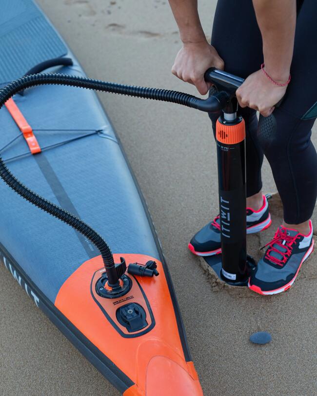 Person pumpt orange-schwarzes Paddleboard auf Sandstrand auf, trägt schwarze Leggings, schwarze Schuhe mit roten Akzenten.
