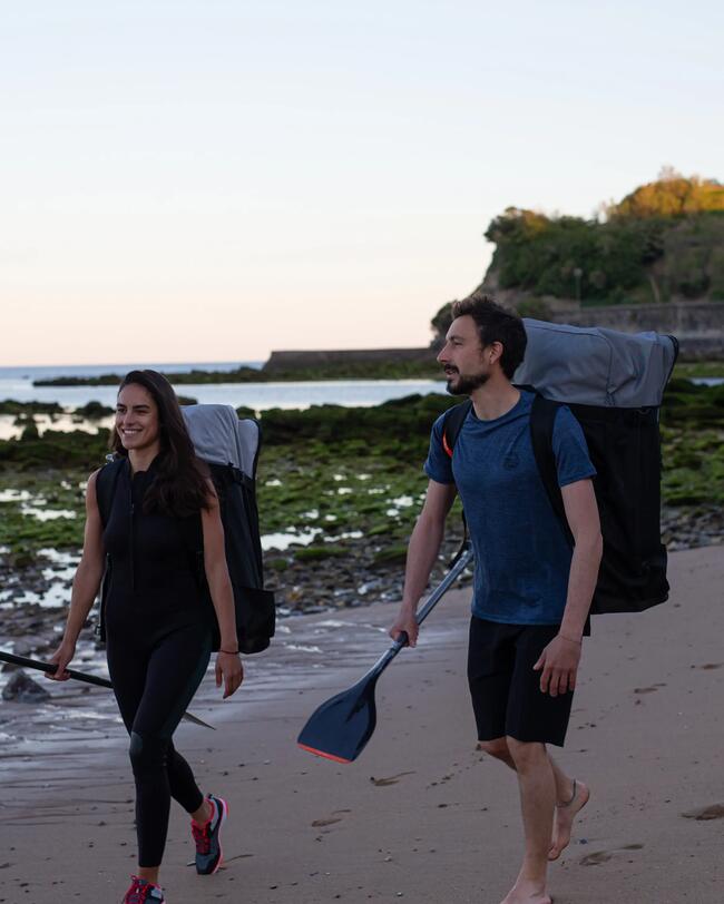 Zwei Personen barfuß am Strand, tragen Kajakausrüstung, ruhige See im Hintergrund, Abendlicht, grüne und sandige Texturen.