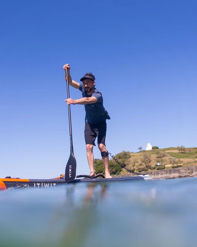 Mann auf Paddleboard, blaues Wasser, klarer Himmel, grüne Küstenlinie, sportliche Haltung.